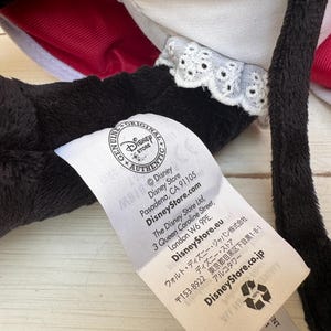 May include: Close-up of a black plush toy with red and white details. A white tag with the Disney Store logo and text is attached. The tag includes the Disney Store website and address in London.