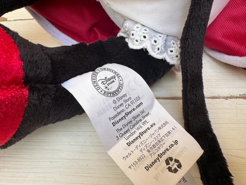 May include: Close-up of a black plush toy with red and white details. A white tag with the Disney Store logo and text is attached. The tag includes the Disney Store website and address in London.
