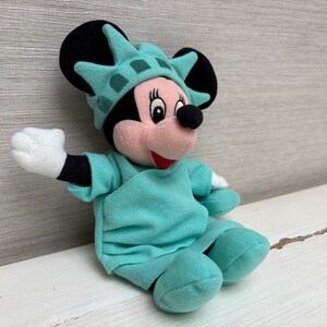 Może przedstawiać: Pluszowa lalka Minnie Mouse przebrana za Statuę Wolności. Lalka ma na sobie turkusowy str&oacute;j i pasującą koronę. Widoczne są czarne uszy i białe rękawiczki Minnie. Lalka siedzi na białej powierzchni.