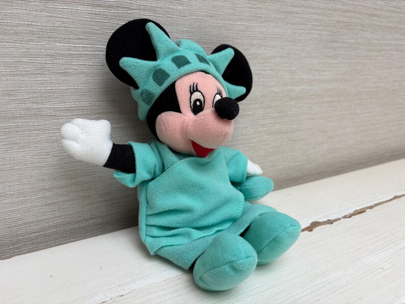 Może przedstawiać: Pluszowa lalka Minnie Mouse przebrana za Statuę Wolności. Lalka ma na sobie turkusowy str&oacute;j i pasującą koronę. Widoczne są czarne uszy i białe rękawiczki Minnie. Lalka siedzi na białej powierzchni.