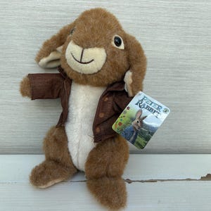 May include: A plush Peter Rabbit toy featuring soft brown fur, a cream-colored belly, and a brown jacket. The rabbit has dark eyes and a stitched smile. A rectangular tag attached to the jacket displays the text Peter Rabbit with an illustration.