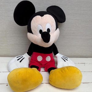 May include: A plush Mickey Mouse toy with large black ears, a black body, red shorts, and yellow shoes. The toy has white gloves with black lines and a smiling face.