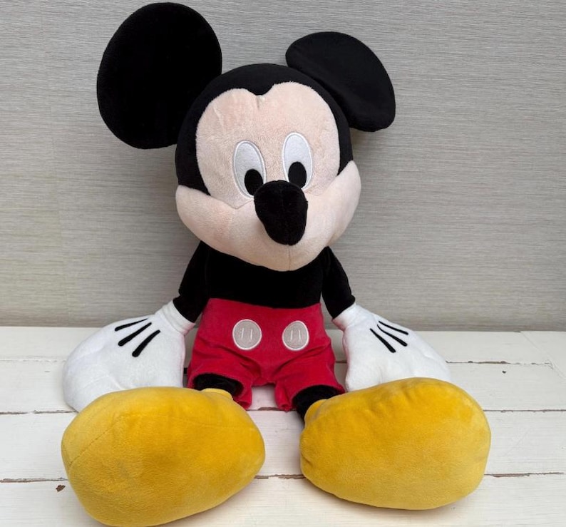 May include: A plush Mickey Mouse toy with large black ears, a black body, red shorts, and yellow shoes. The toy has white gloves with black lines and a smiling face.