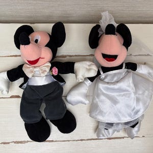 May include: Two plush Mickey and Minnie Mouse wedding dolls. Mickey is dressed in a black tuxedo with a white bow tie and a pink rose. Minnie wears a white satin wedding dress and veil. Both have black ears and red smiles.