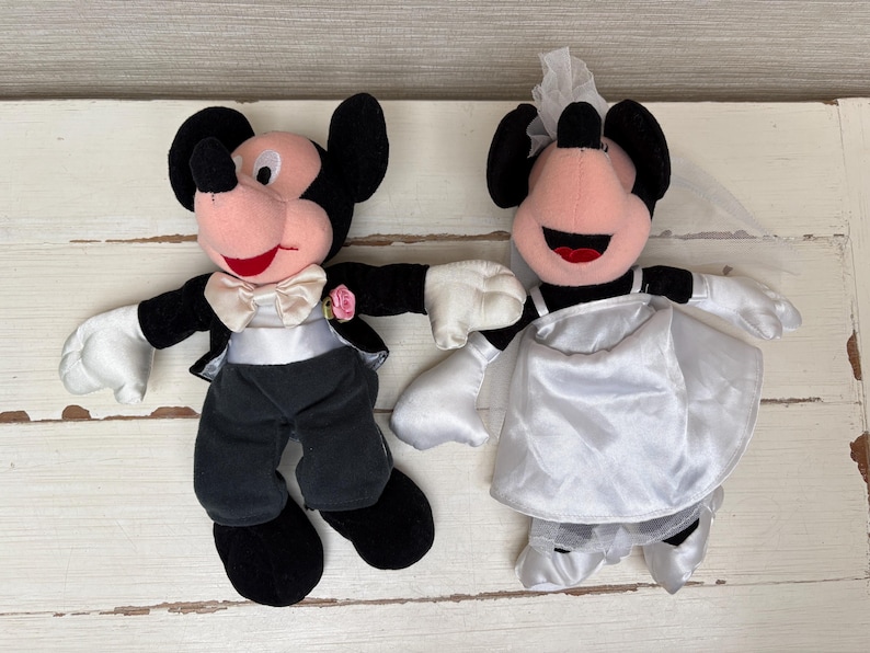 May include: Two plush Mickey and Minnie Mouse wedding dolls. Mickey is dressed in a black tuxedo with a white bow tie and a pink rose. Minnie wears a white satin wedding dress and veil. Both have black ears and red smiles.