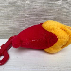 May include: A plush toy in the shape of a hand, with a red body and yellow fingers. A red plastic clip is attached to the toy. The toy is lying on a white surface.