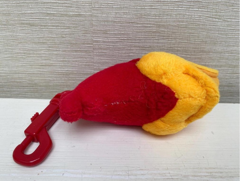 May include: A plush toy in the shape of a hand, with a red body and yellow fingers. A red plastic clip is attached to the toy. The toy is lying on a white surface.