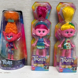 Puede incluir: Tres muñecas Trolls en su embalaje original. Las muñecas tienen piel rosa, ojos grandes y pelo colorido. Una muñeca tiene pelo naranja, mientras que las otras dos tienen pelo rosa y amarillo. El embalaje presenta los logotipos "Trolls World Tour" y "Trolls Band Together".