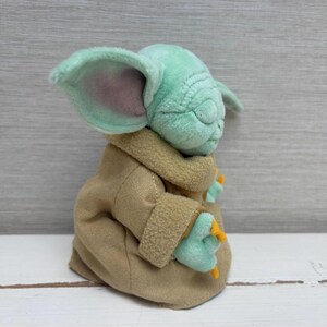 May include: A soft toy of a character with large ears, green skin, and a tan robe. The toy has closed eyes and small hands with orange claws. The character is from a popular science fiction franchise.