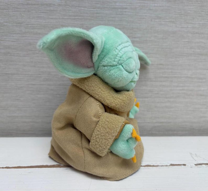 May include: A soft toy of a character with large ears, green skin, and a tan robe. The toy has closed eyes and small hands with orange claws. The character is from a popular science fiction franchise.