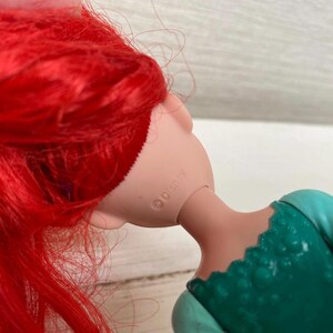 Op de afbeelding: Een pop met felrood haar en een zeegroene jurk. De nek van de pop is zichtbaar, met het woord "Disney" erop gedrukt. Het haar van de pop is lang en vloeiend en de jurk heeft een getextureerd lijfje.