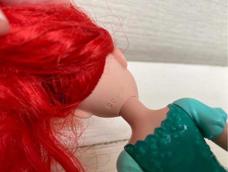 Op de afbeelding: Een pop met felrood haar en een zeegroene jurk. De nek van de pop is zichtbaar, met het woord "Disney" erop gedrukt. Het haar van de pop is lang en vloeiend en de jurk heeft een getextureerd lijfje.