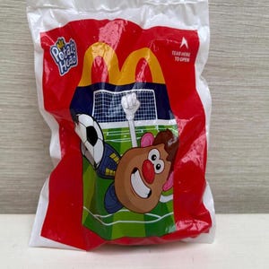 Pode incluir: Saco plástico vermelho e branco selado com um personagem do Mr. Potato Head jogando futebol. A sacola tem o logotipo dos arcos dourados do McDonald's e as palavras "Mr. Potato Head" impressas. A sacola também tem as palavras "Rasgue aqui para abrir".