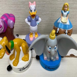 Könnte beinhalten: Eine Sammlung von Disney-Charakterfiguren, darunter Pluto, Dumbo, Daisy Duck, Alice im Wunderland und die Grinsekatze. Jede Figur ist farbenfroh und auf einem Sockel mit dem Disneyland-Logo montiert.