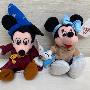 May include: Two Disney plush toys: Mickey Mouse dressed as a sorcerer with a blue hat and Minnie Mouse with a blue bow. Both have tags from The Disney Store. The Mickey Mouse plush is wearing a burgundy robe and brown shoes.