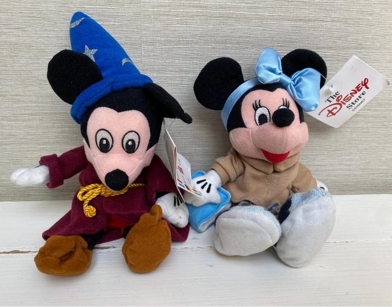 May include: Two Disney plush toys: Mickey Mouse dressed as a sorcerer with a blue hat and Minnie Mouse with a blue bow. Both have tags from The Disney Store. The Mickey Mouse plush is wearing a burgundy robe and brown shoes.