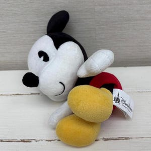 Op de afbeelding: Een pluche Mickey Mouse-speelgoed in een zijliggende pose. Het speelgoed heeft een wit gezicht en lichaam, zwarte oren en gele voeten. Het heeft een rode broek en witte handschoenen. Een Disney Parks-label is bevestigd.