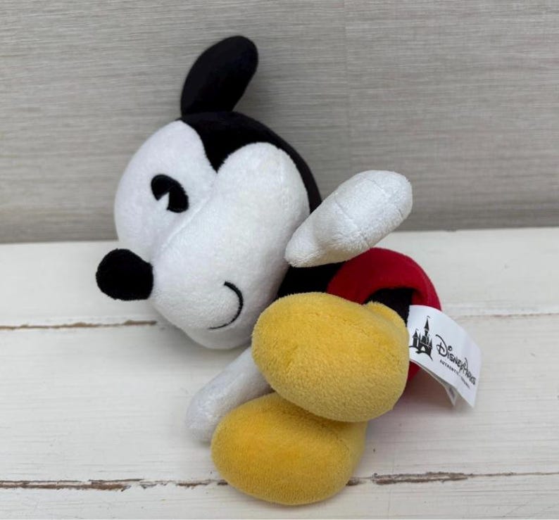 Op de afbeelding: Een pluche Mickey Mouse-speelgoed in een zijliggende pose. Het speelgoed heeft een wit gezicht en lichaam, zwarte oren en gele voeten. Het heeft een rode broek en witte handschoenen. Een Disney Parks-label is bevestigd.