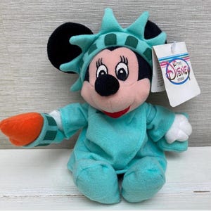 May include: A plush Minnie Mouse doll dressed as the Statue of Liberty. The doll wears a turquoise robe and crown, with an orange torch. The tag reads "The Disney Store".