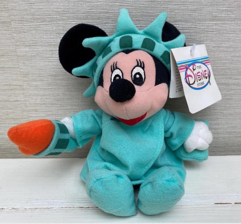 May include: A plush Minnie Mouse doll dressed as the Statue of Liberty. The doll wears a turquoise robe and crown, with an orange torch. The tag reads "The Disney Store".