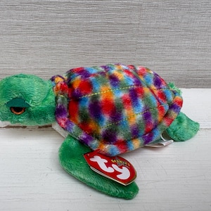 May include: A plush turtle toy with a green head, flippers, and a colorful shell. The shell features a pattern of red, blue, yellow, and purple. A red tag with white text is attached to the turtle's flipper.