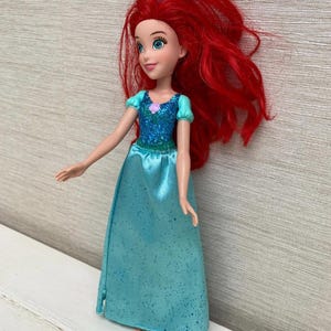 Op de afbeelding: Pop van een personage met lang, felrood haar. De pop draagt een turquoise jurk met een sprankelend lijfje en een kleine paarse bloem. De pop heeft blauwe ogen en een zachte uitdrukking.