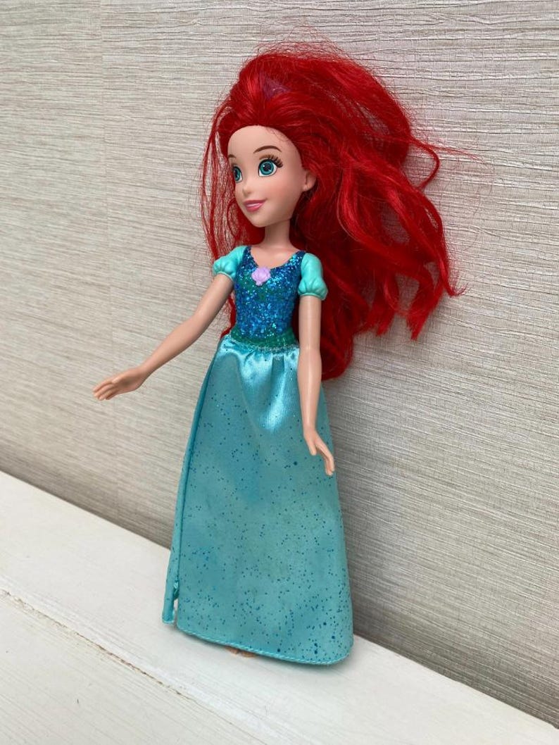 Op de afbeelding: Pop van een personage met lang, felrood haar. De pop draagt een turquoise jurk met een sprankelend lijfje en een kleine paarse bloem. De pop heeft blauwe ogen en een zachte uitdrukking.