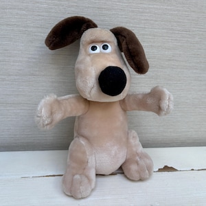 May include: A plush dog toy featuring a light tan body, two dark brown floppy ears, and a prominent black nose. The character has large white eyes with black pupils, a soft textured surface, and outstretched arms in a standing position.