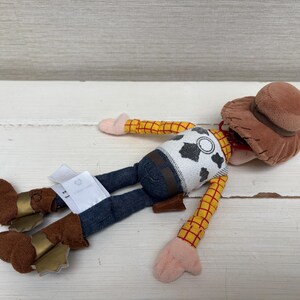 May include: A plush Woody doll from the Toy Story franchise. The doll is wearing a brown cowboy hat, a yellow plaid shirt, a white vest with black cow spots, blue jeans, and brown boots with gold accents. The doll is lying on a white surface.