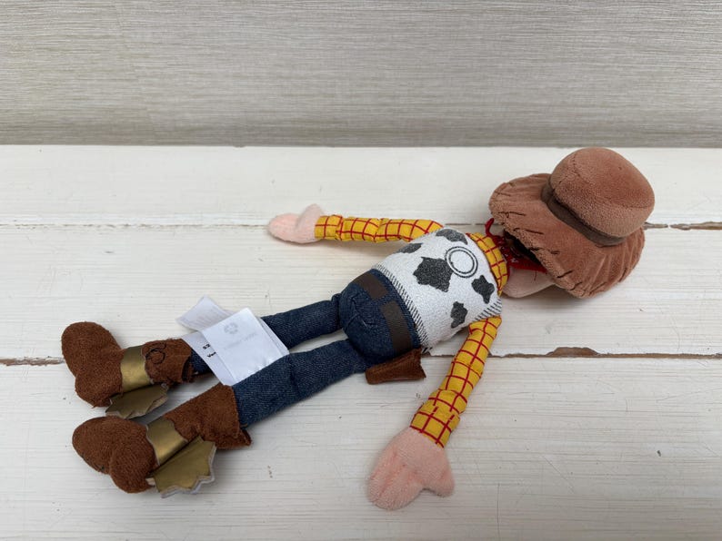 May include: A plush Woody doll from the Toy Story franchise. The doll is wearing a brown cowboy hat, a yellow plaid shirt, a white vest with black cow spots, blue jeans, and brown boots with gold accents. The doll is lying on a white surface.