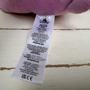 Puede incluir: Un peluche morado de un personaje de Disney con una etiqueta blanca adjunta. La etiqueta incluye el logotipo de Disney y texto en varios idiomas. El juguete descansa sobre una superficie de madera clara y desgastada.