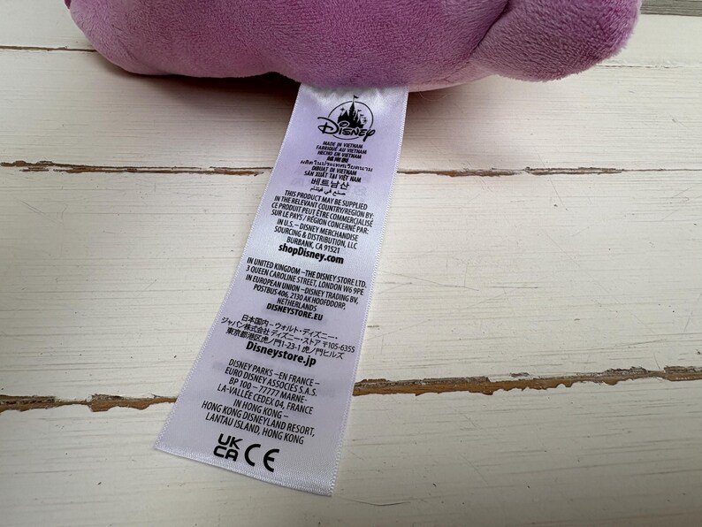 Puede incluir: Un peluche morado de un personaje de Disney con una etiqueta blanca adjunta. La etiqueta incluye el logotipo de Disney y texto en varios idiomas. El juguete descansa sobre una superficie de madera clara y desgastada.