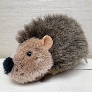 May include: A plush hedgehog toy featuring a soft tan face with a small black nose and dark eyes. The back is covered in long, shaggy brown and gray faux fur, and the ears are made of a smooth, light tan felt material. The toy has a rounded shape.