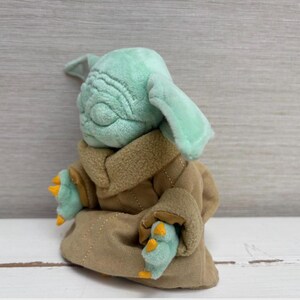 May include: A plush toy of a character with large ears, green skin, and a tan robe. The toy has a detailed face with wrinkles and is seated. The character's hands and feet are visible, with yellow claws.