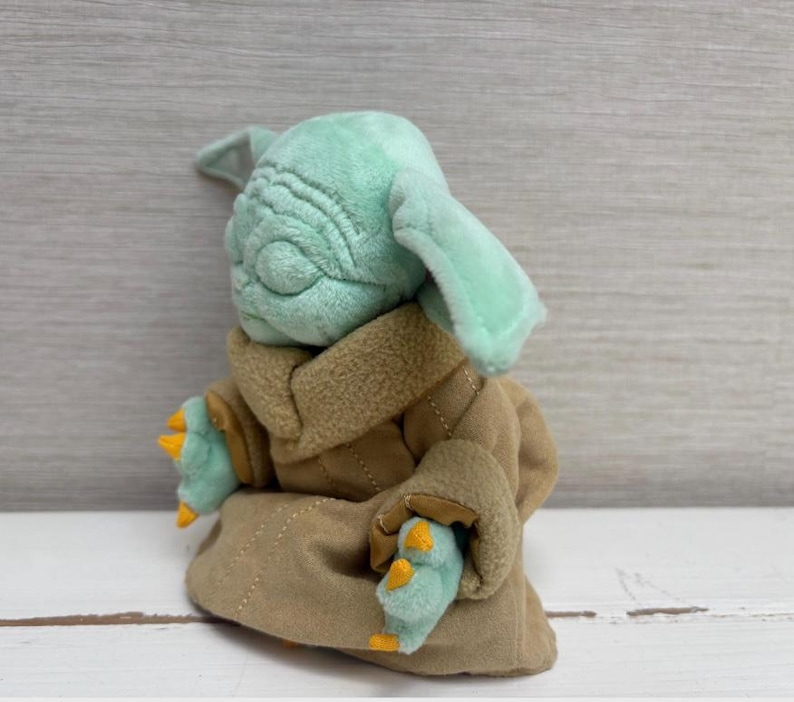 May include: A plush toy of a character with large ears, green skin, and a tan robe. The toy has a detailed face with wrinkles and is seated. The character's hands and feet are visible, with yellow claws.