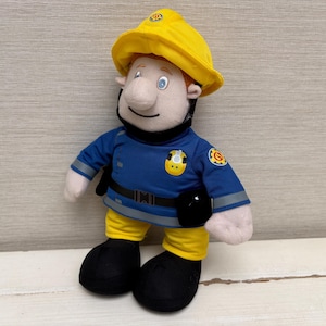 Può includere: Una bambola di peluche pompiere con casco giallo, giacca blu e pantaloni gialli. La bambola ha una cintura nera, stivali neri e un accessorio nero. Il viso e le braccia sono bianche.