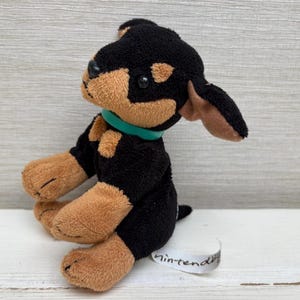 May include: A plush toy dog in black and tan with a green collar. The stuffed animal has a small tail and a tag that says "Nintendo". The dog is sitting on a white surface.