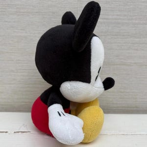 Op de afbeelding: Een pluche Mickey Mouse-speelgoed, met een zwarte kop en oren, een wit gezicht, een rode broek, gele schoenen en witte handschoenen. Het knuffeldier zit, met zijn zijprofiel zichtbaar. Het speelgoed is gemaakt van zachte stof.
