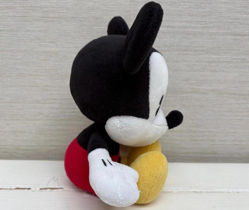 Op de afbeelding: Een pluche Mickey Mouse-speelgoed, met een zwarte kop en oren, een wit gezicht, een rode broek, gele schoenen en witte handschoenen. Het knuffeldier zit, met zijn zijprofiel zichtbaar. Het speelgoed is gemaakt van zachte stof.