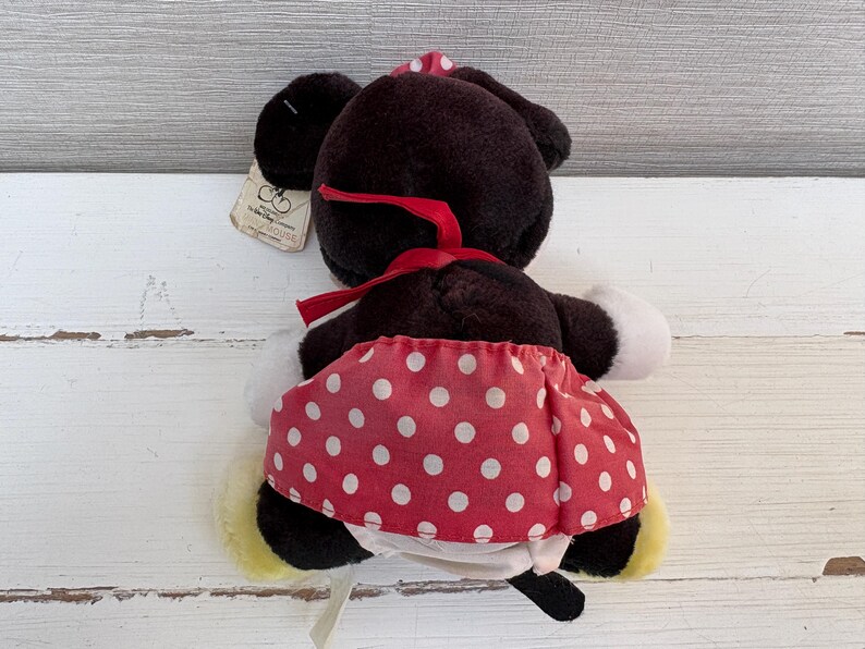 K&ouml;nnte beinhalten: Eine Pl&uuml;schpuppe von Minnie Mouse, von hinten gesehen, hat einen schwarzen Kopf und Ohren, eine rote Schleife und einen rot-wei&szlig; gepunkteten Rock. Die Puppe hat wei&szlig;e Handschuhe und gelbe Schuhe. Ein Etikett ist angebracht.