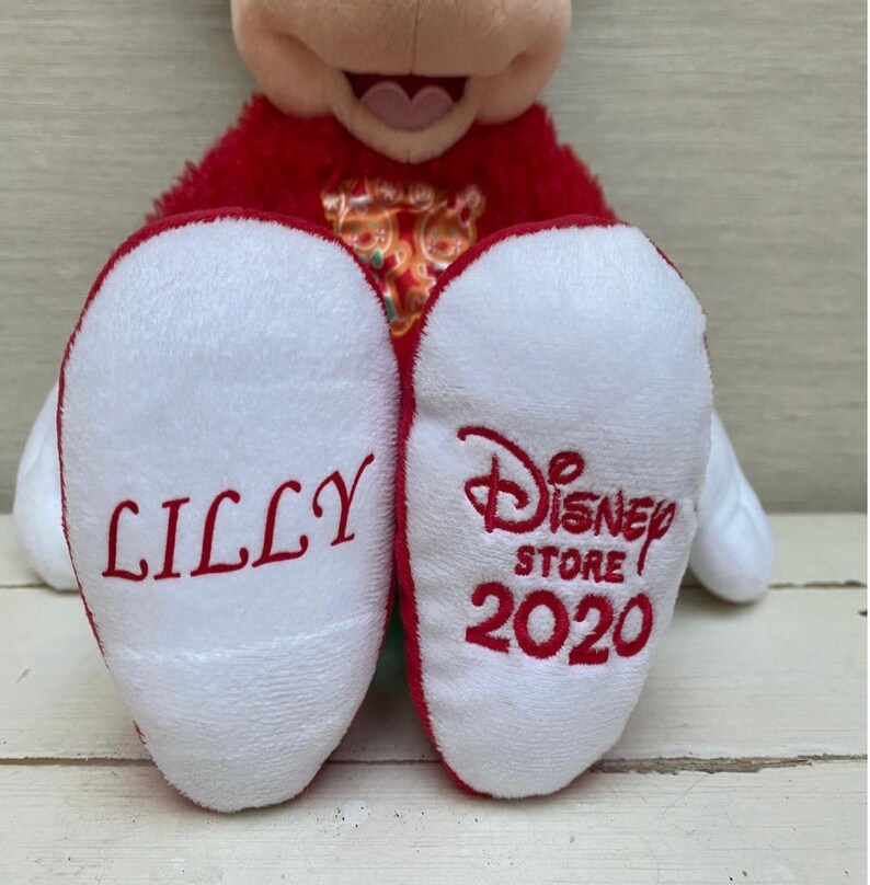 May include: A plush Mickey Mouse toy with red and white accents. The soles of the feet are white with red embroidered text that reads "LILLY" on one foot and "Disney Store 2020" on the other.