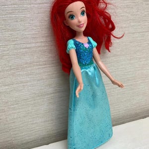 Op de afbeelding: Een pop van een personage met felrood haar, blauwe ogen en een glimlachende uitdrukking. De pop draagt een turquoise jurk met een sprankelend lijfje en mouwen. Een kleine roze bloem siert het lijfje. De pop staat op een wit oppervlak.