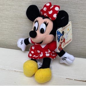 May include: A Minnie Mouse soft toy, dressed in a red and white polka dot dress and bow. The toy has black ears, a black nose, and white gloves. A Disneyland tag is attached.