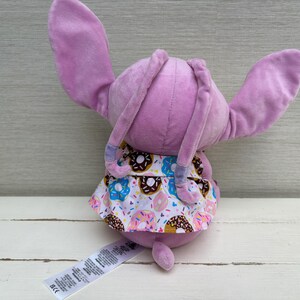 Puede incluir: Un peluche de conejo lavanda con orejas largas y una cola peque&ntilde;a. El conejo lleva una falda estampada con coloridos donuts. Una etiqueta blanca con texto est&aacute; adherida al cuerpo del conejo.