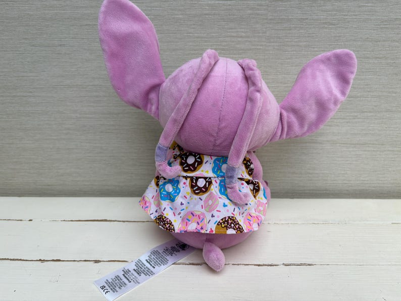 Puede incluir: Un peluche de conejo lavanda con orejas largas y una cola peque&ntilde;a. El conejo lleva una falda estampada con coloridos donuts. Una etiqueta blanca con texto est&aacute; adherida al cuerpo del conejo.