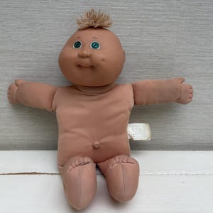 May include: A vintage Cabbage Patch Kid doll with a tan skin tone, green eyes, and a tuft of light brown hair. The doll is wearing a matching tan outfit and has its arms outstretched. The doll is sitting on a white surface.