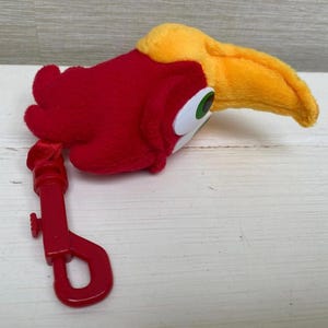 May include: A plush toucan head with a red body, yellow beak, and green and white eyes. A red plastic clip is attached to the toucan's body. The toy is designed to be a keychain or bag charm.