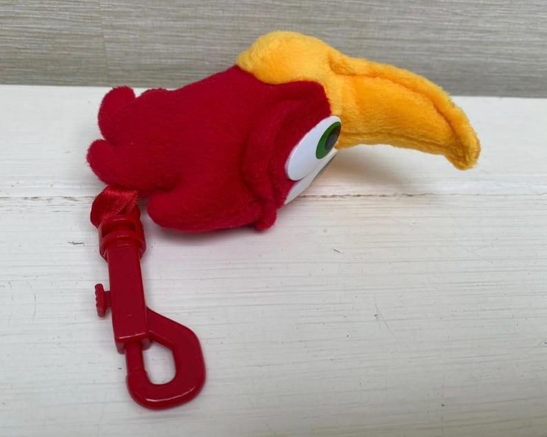 May include: A plush toucan head with a red body, yellow beak, and green and white eyes. A red plastic clip is attached to the toucan's body. The toy is designed to be a keychain or bag charm.