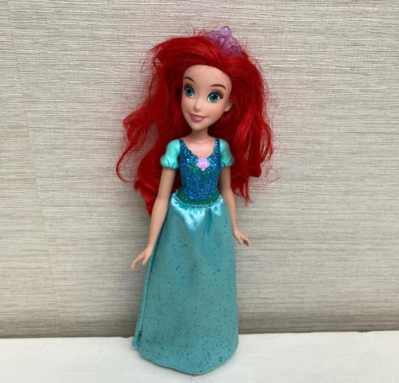 Op de afbeelding: Pop van een personage met felrood haar, gekleed in een turquoise jurk met een sprankelend lijfje en een paarse bloemaccent. De pop heeft blauwe ogen en een tiara. De pop is een populair personage uit een animatiefilm voor kinderen.