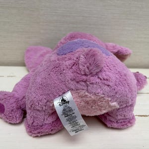 May include: A plush toy in shades of purple, with a darker purple patch on its back. The toy has a soft, furry texture and a Disney tag attached. The toy is lying on a light-colored wooden surface.